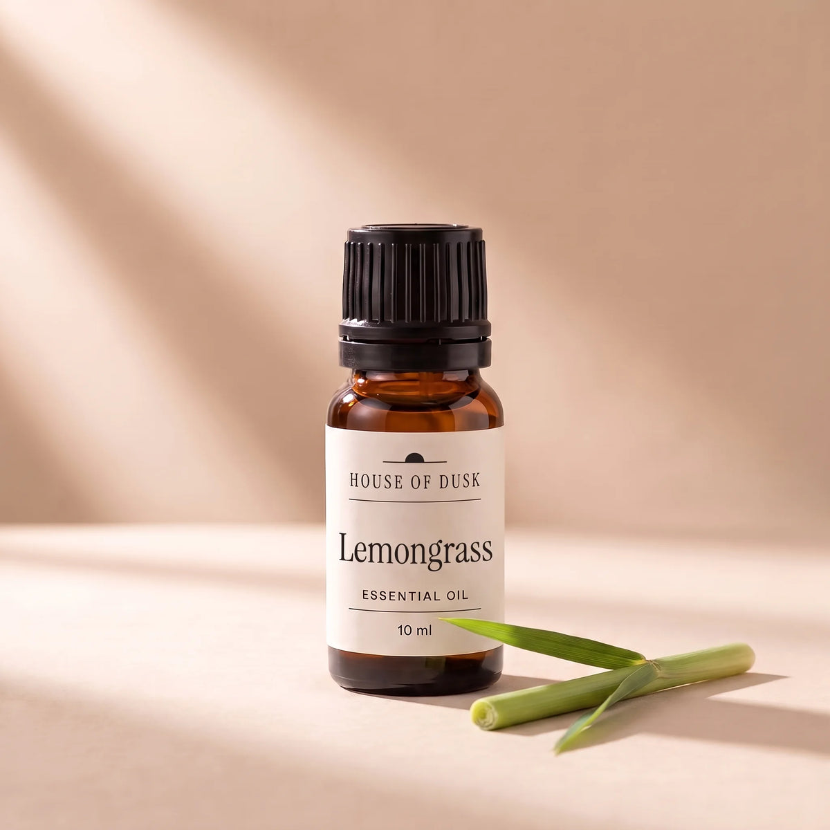 Lemongrass essential oil in a 10 ml amber glass bottle with a House of Dusk label, beside a short piece of fresh lemongrass stalk with one leaf blade