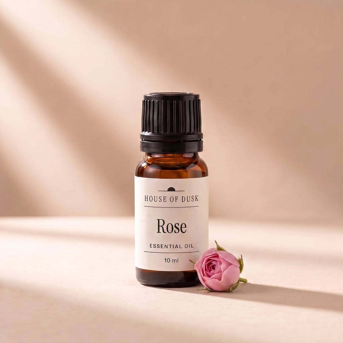 Rose essential oil in a 10 ml amber glass bottle with a House of Dusk label, beside one small pink damask rose bud