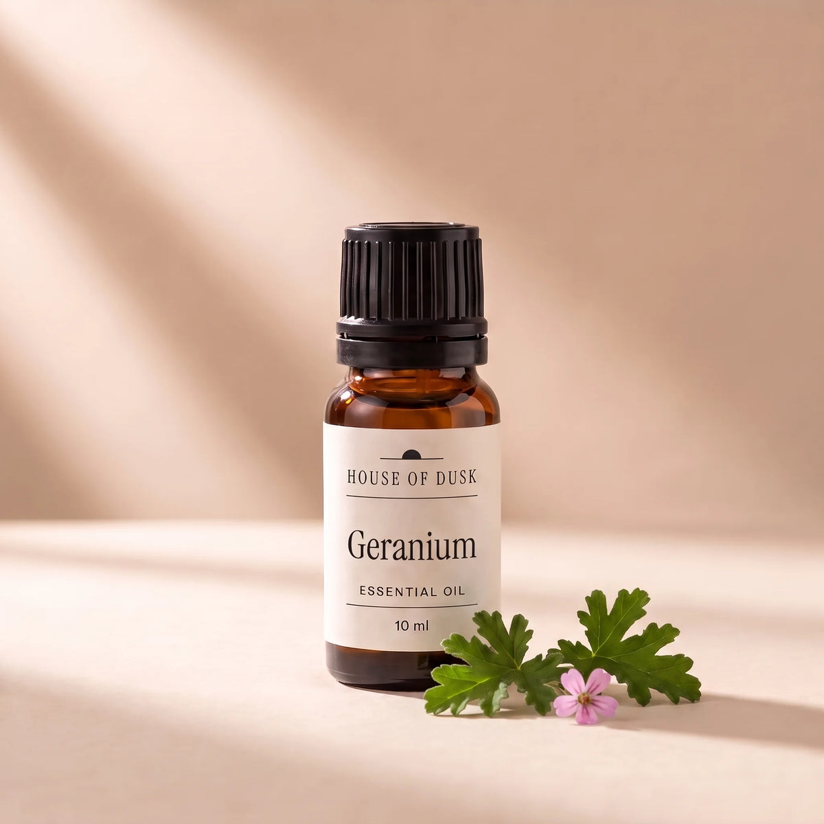 Geranium essential oil in a 10 ml amber glass bottle with a House of Dusk label, beside two deeply lobed rose geranium leaves with one tiny pink flower