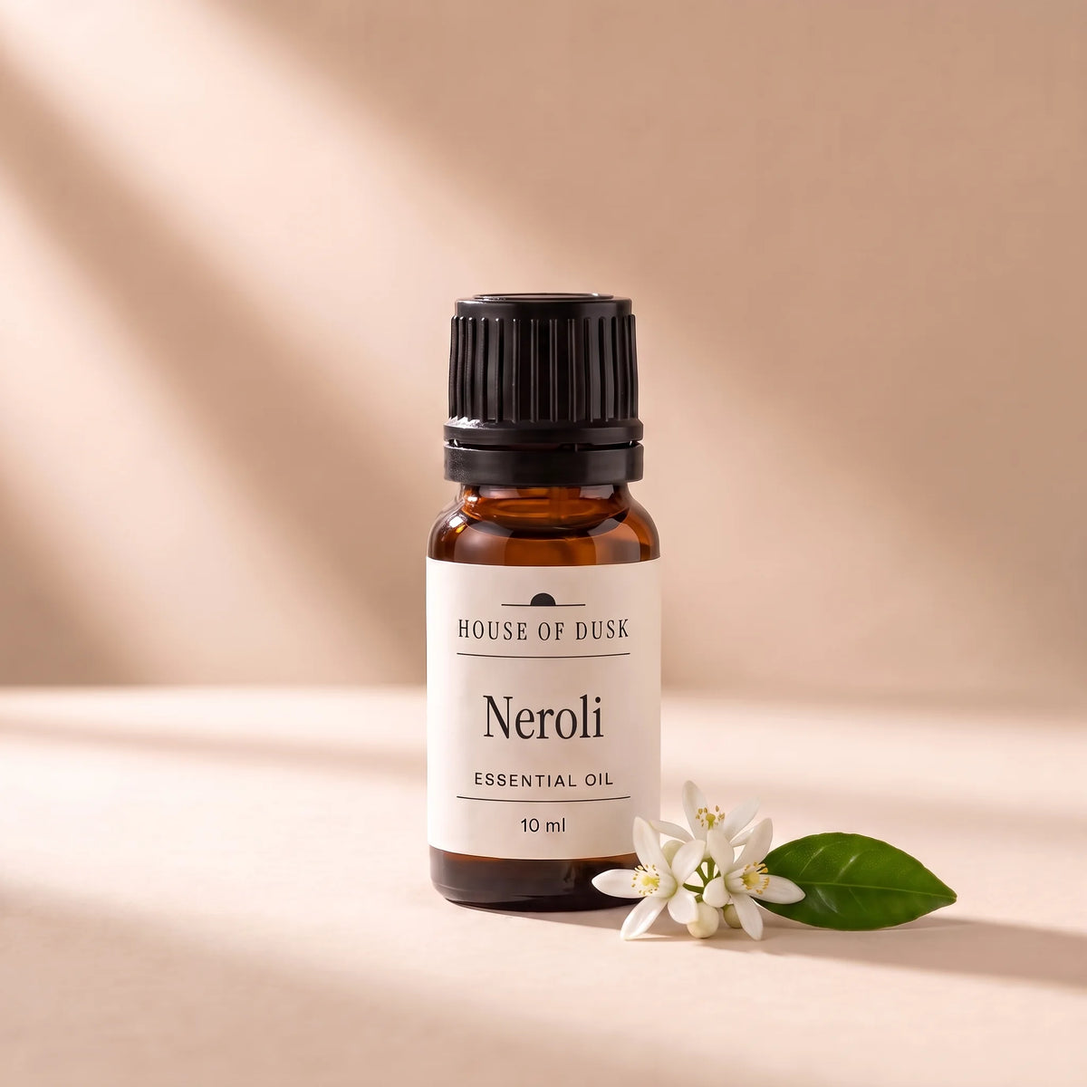 Neroli essential oil in a 10 ml amber glass bottle with a House of Dusk label, beside a small sprig of white orange blossom flowers with one glossy leaf