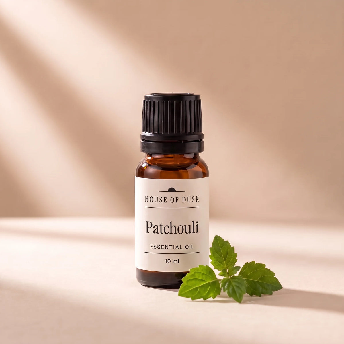 Patchouli essential oil in a 10 ml amber glass bottle with a House of Dusk label, beside a small sprig of green patchouli leaves