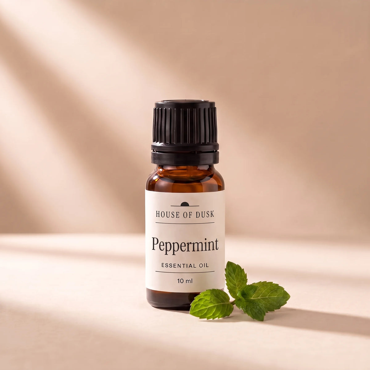 Peppermint essential oil in a 10 ml amber glass bottle with a House of Dusk label, beside a small sprig of fresh peppermint with a few green leaves