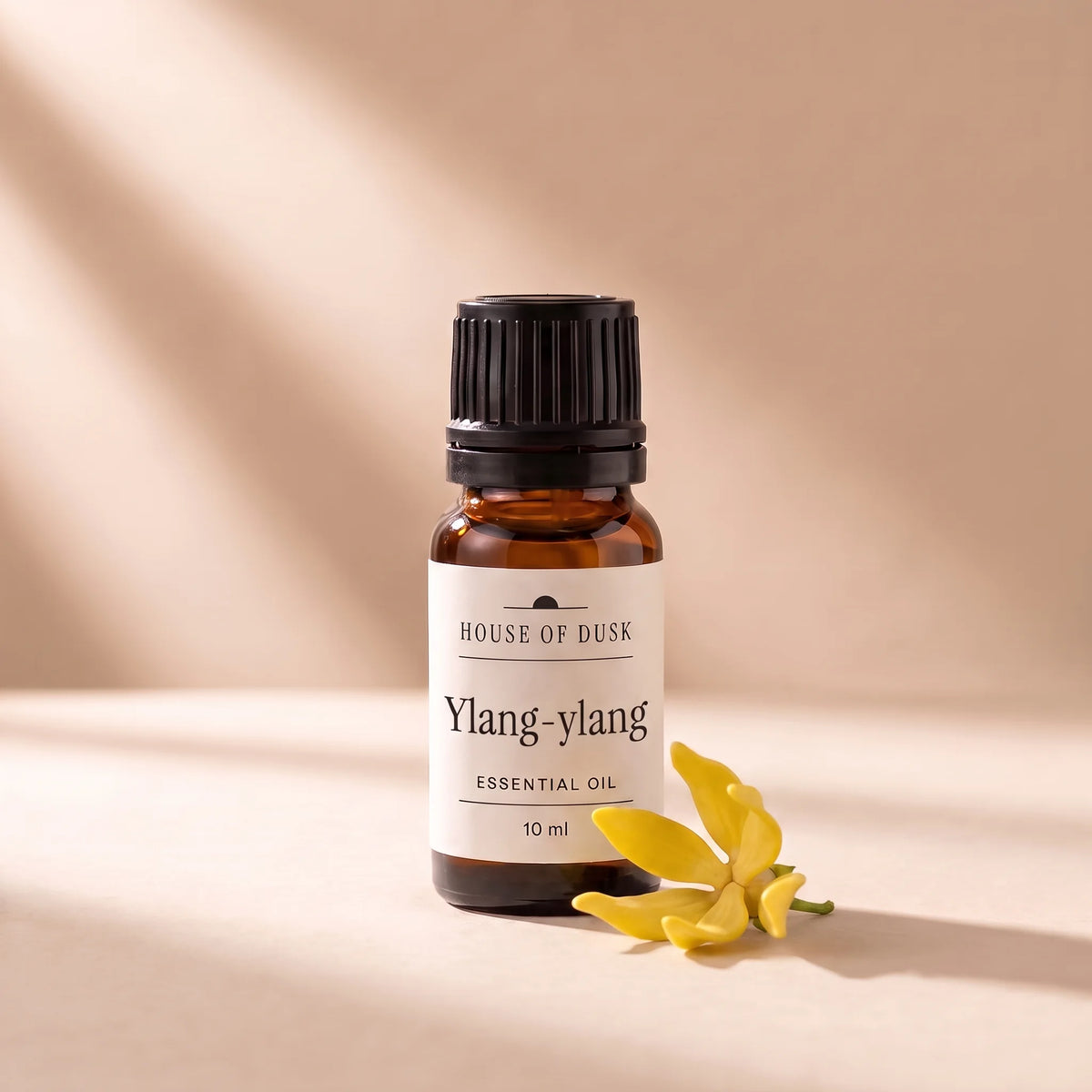 Ylang-ylang essential oil in a 10 ml amber glass bottle with a House of Dusk label, beside one yellow ylang ylang flower with long drooping petals