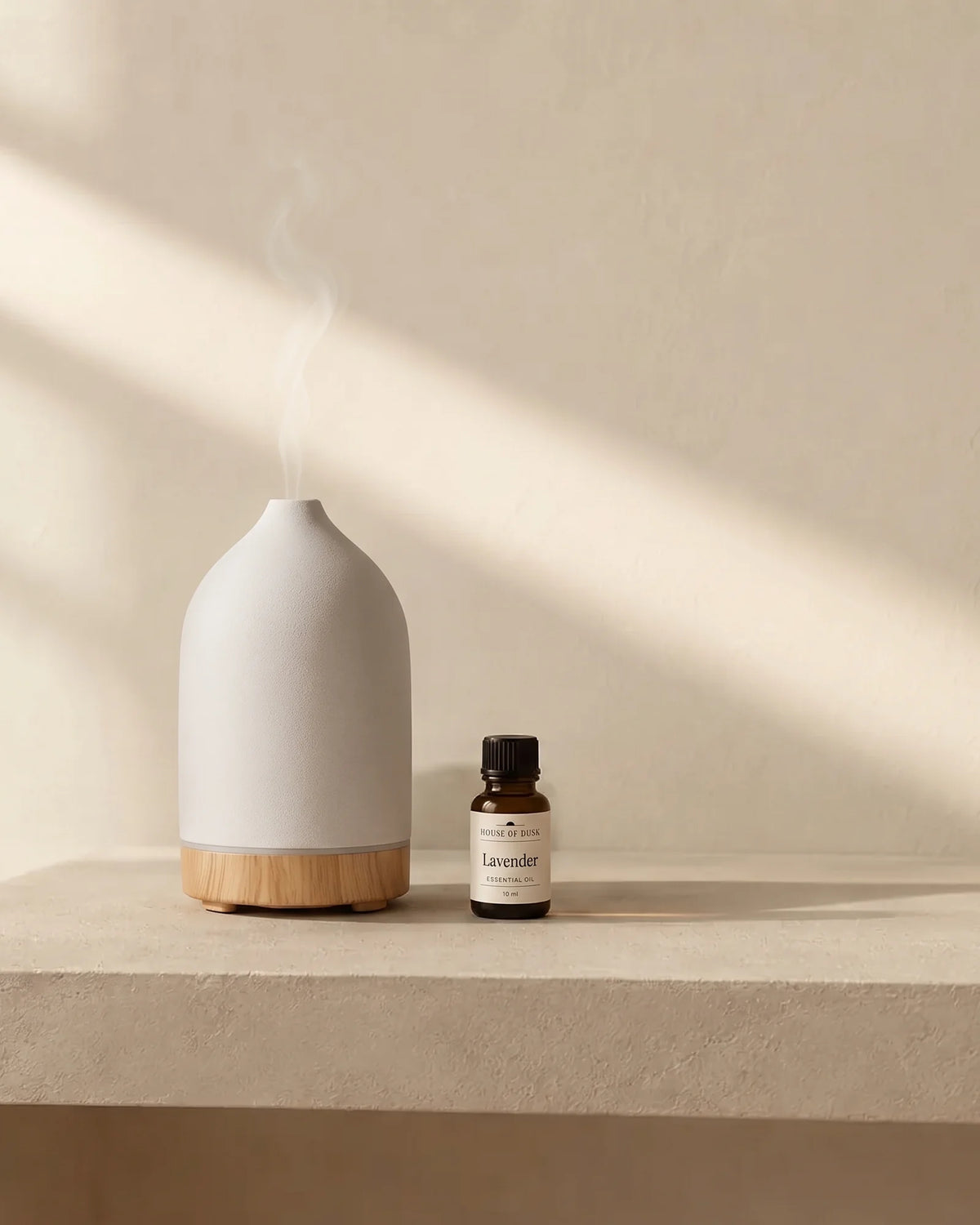 White matte ceramic diffuser on a wood grain base and a 10 ml House of Dusk lavender essential oil bottle on a plaster console